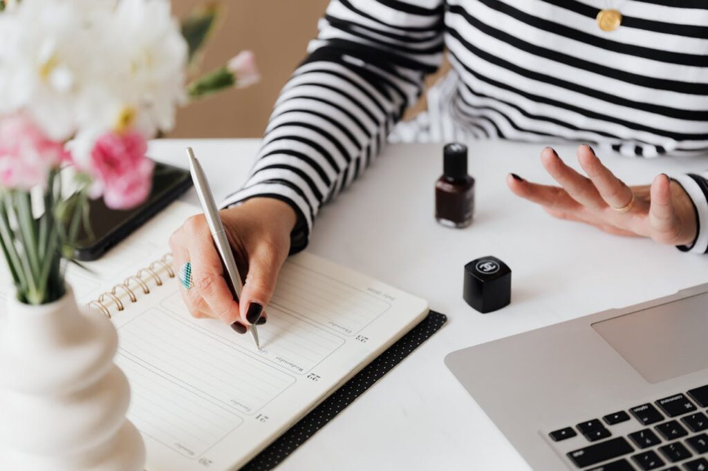 Unrecognizable female using pen and writing notes in notepad while sitting at table and waiting when polish nail getting dry